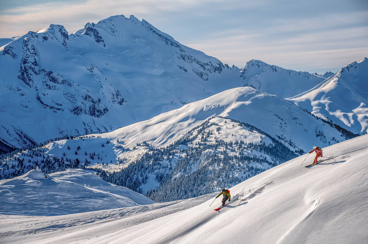 two skiers descend through the powder on a steep slope, several mountain peaks behind them