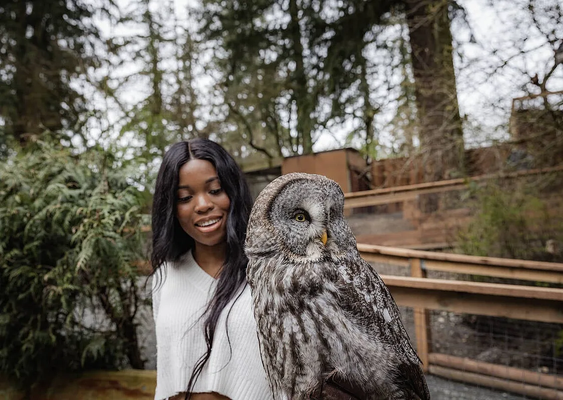 A woman holds an owl at The Raptors near Duncan. They are outside at an enclosure with walkways and trees surrounding them.