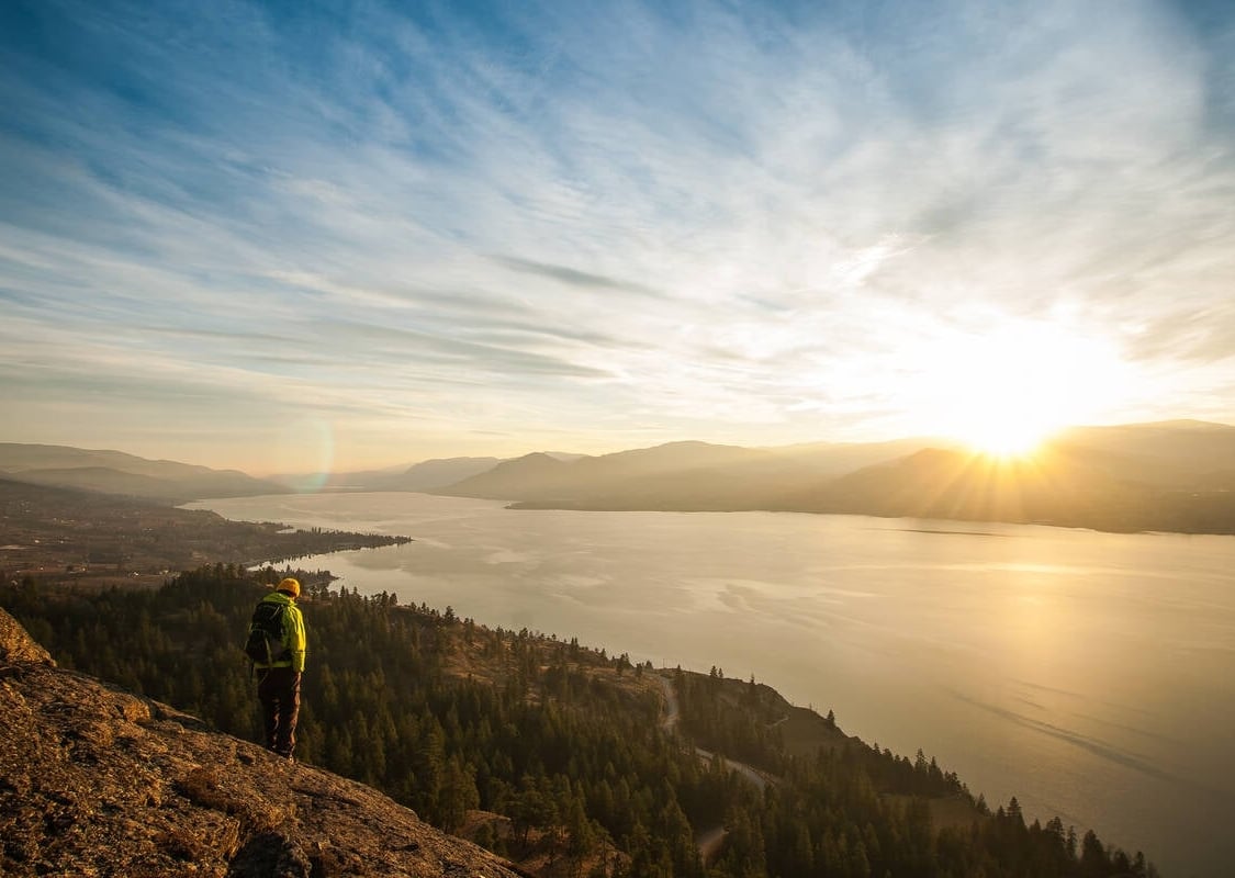 A person stands on a rocky bluff overlooking the lush Okanagan Valley and the large lake as the sun sets on the horizon