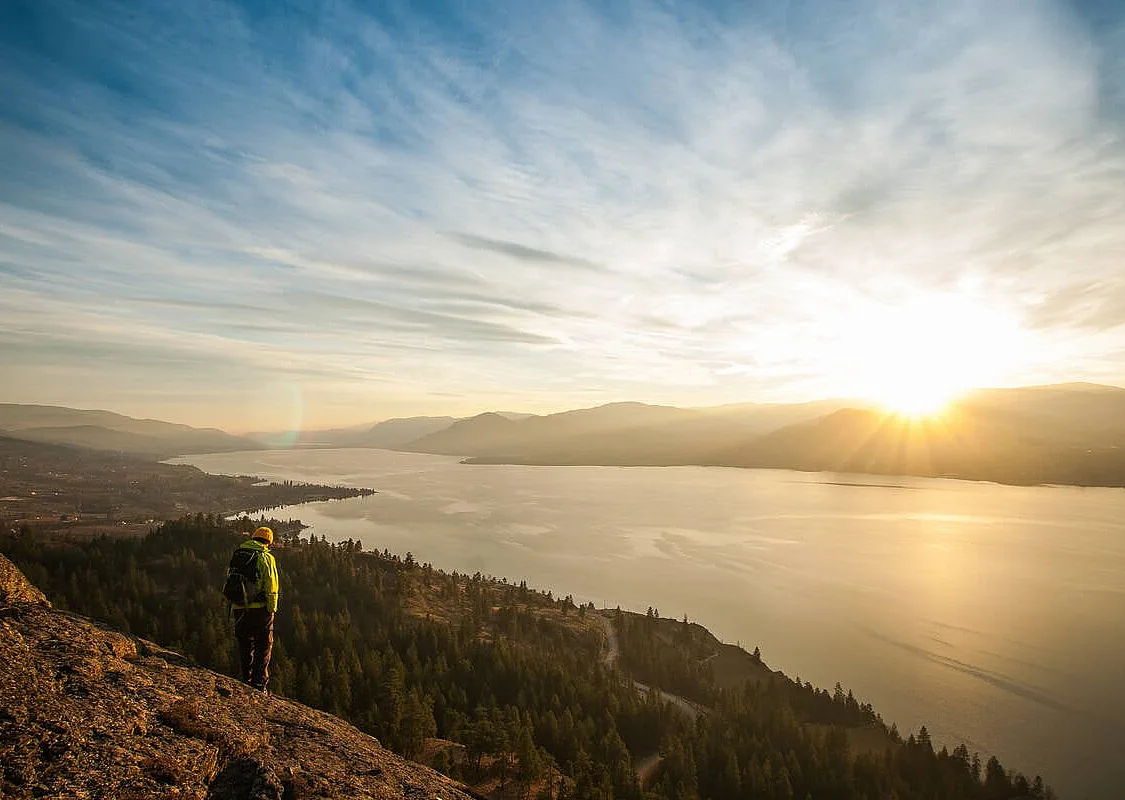 A person stands on a rocky bluff overlooking the lush Okanagan Valley and the large lake as the sun sets on the horizon