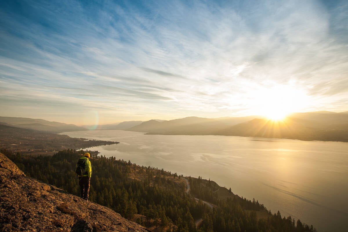 A person stands on a rocky bluff overlooking the lush Okanagan Valley and the large lake as the sun sets on the horizon