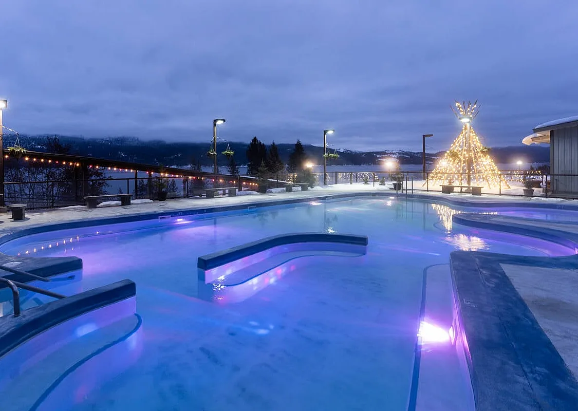 The rounded edges of an empty hot springs pool at dusk. Lights illuminate a teepee structure at the far end of the pool, and in the distance there is a lake and some trees. The sky is cloudy.