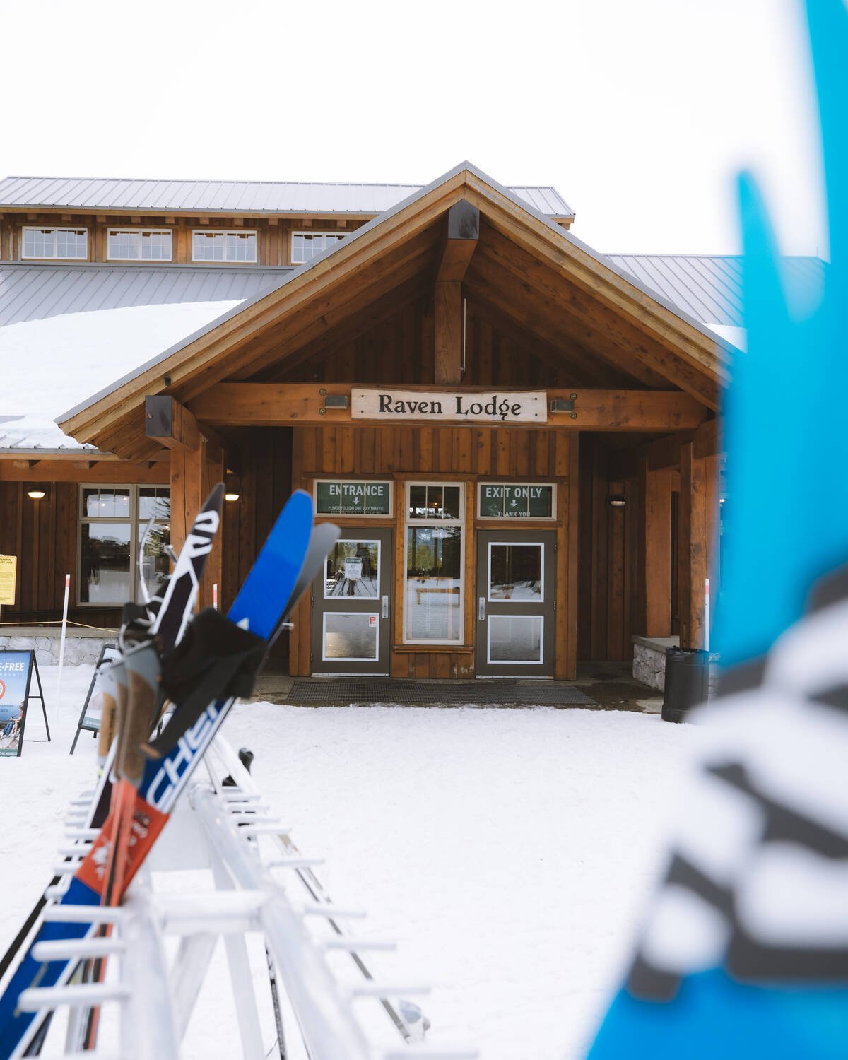 A wooden lodge with snow in the roof and a metal rack with skis on the left of the frame