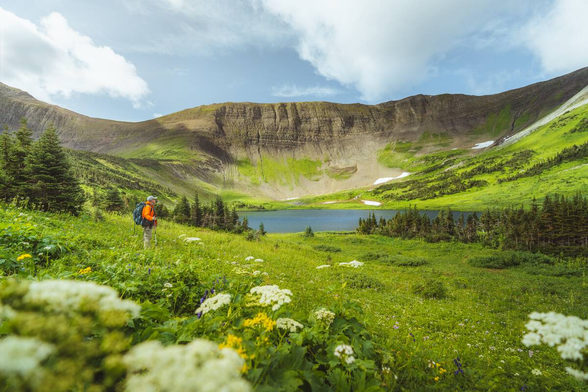 A hiker wearing an orange jacket stands in a lush, green alpine meadow with a small lake and mountains in the backdrop.