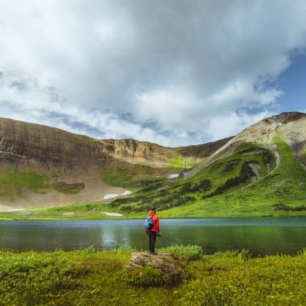 A person stands on a rock overlooking a small alpine lake, surrounded by lush green alpine foliage and rocky mountaintops.