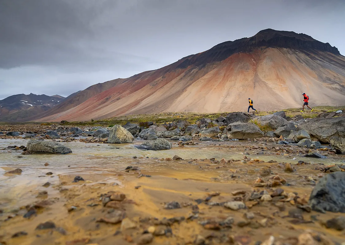 Two trail runners look tiny against the backdrop of a towering Mt. Edziza,streaked with rust and bone colours.