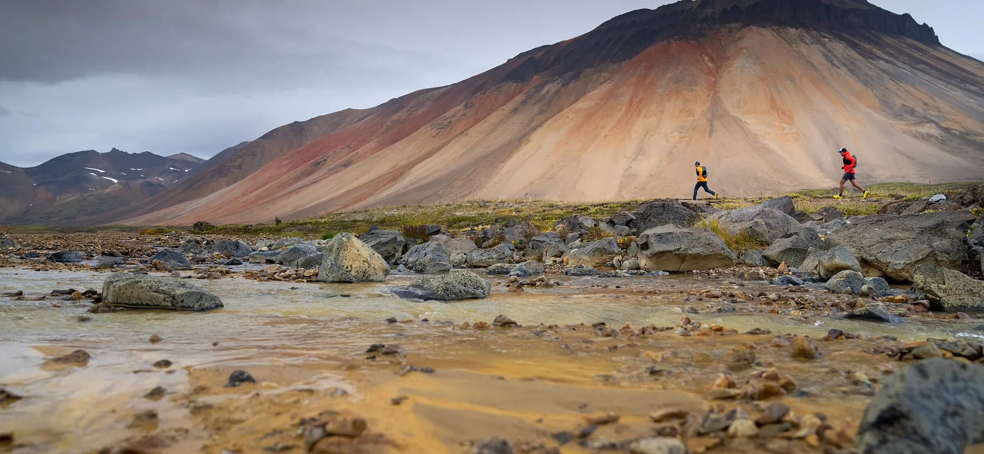 Two trail runners look tiny against the backdrop of a towering Mt. Edziza,streaked with rust and bone colours.