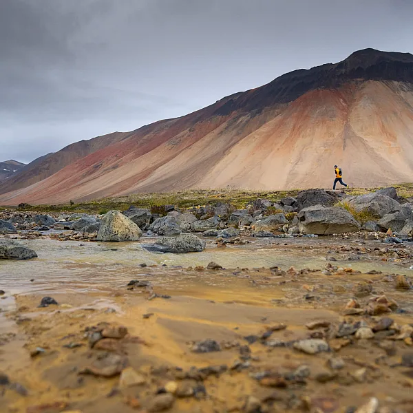 Two trail runners look tiny against the backdrop of a towering Mt. Edziza,streaked with rust and bone colours.