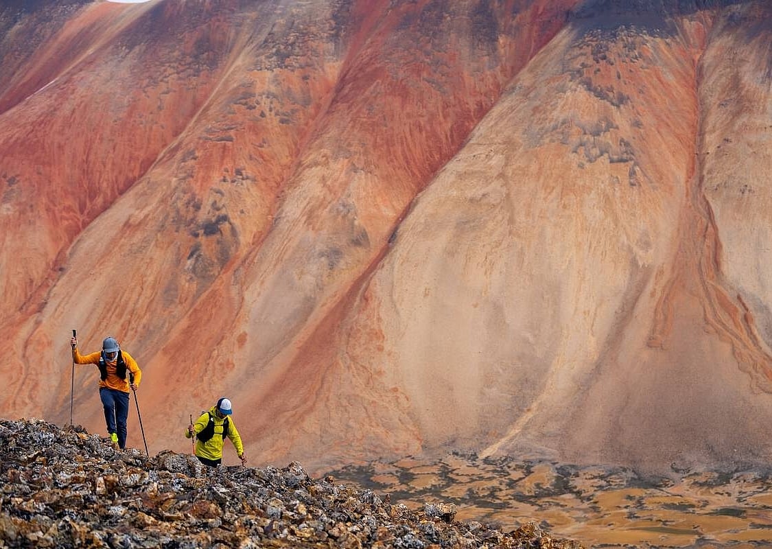 Zwei Personen wandern mit Wanderstöcken vor einem rot und orange gestreiften Berg im Hintergrund.