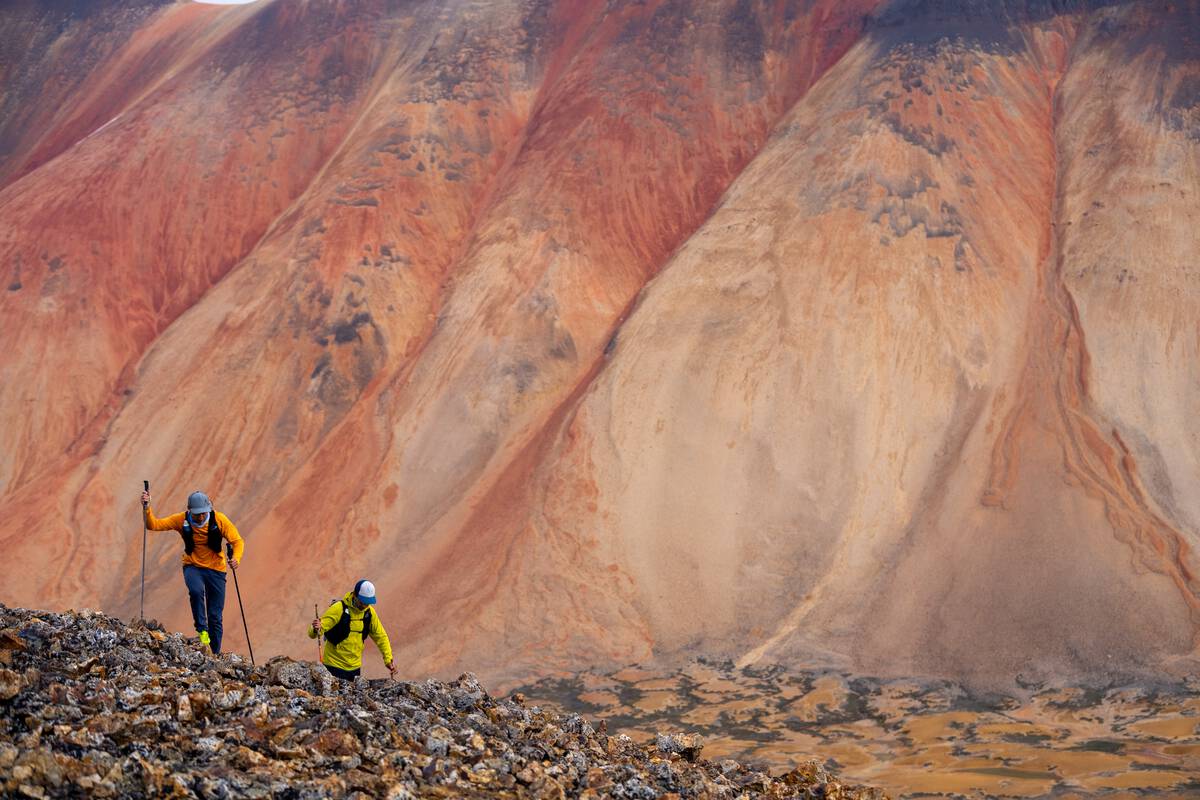 Two people with hiking poles hike with red and orange streaked mountain in the background.