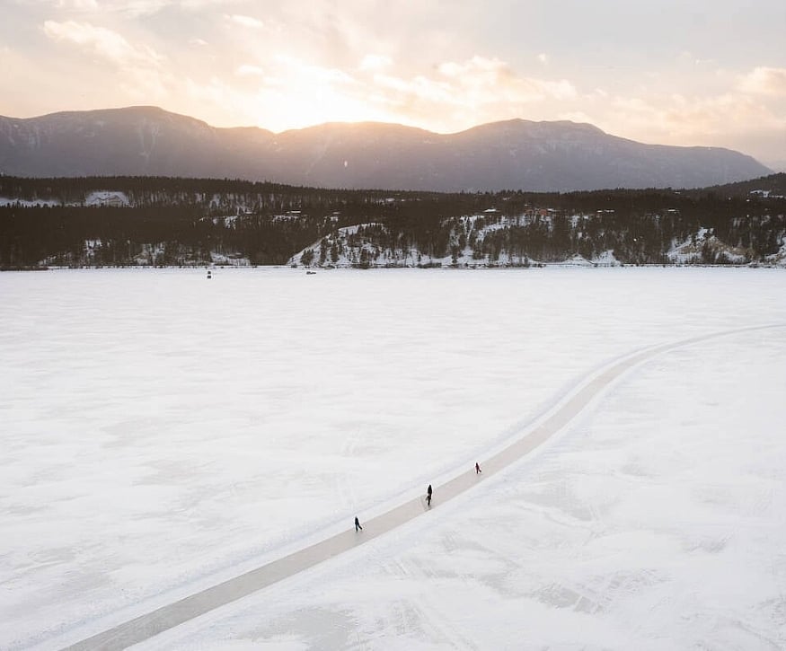 A wavy, icy surface runs through a field of snow with three ice skaters. In the background are tree-covered hills and the sun setting behind distant mountains.