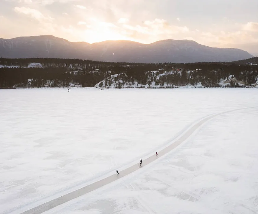 Una superficie ondulada y helada atraviesa un campo de nieve con tres patinadores sobre hielo. Al fondo hay colinas cubiertas de árboles y la puesta de sol detrás de montañas distantes.