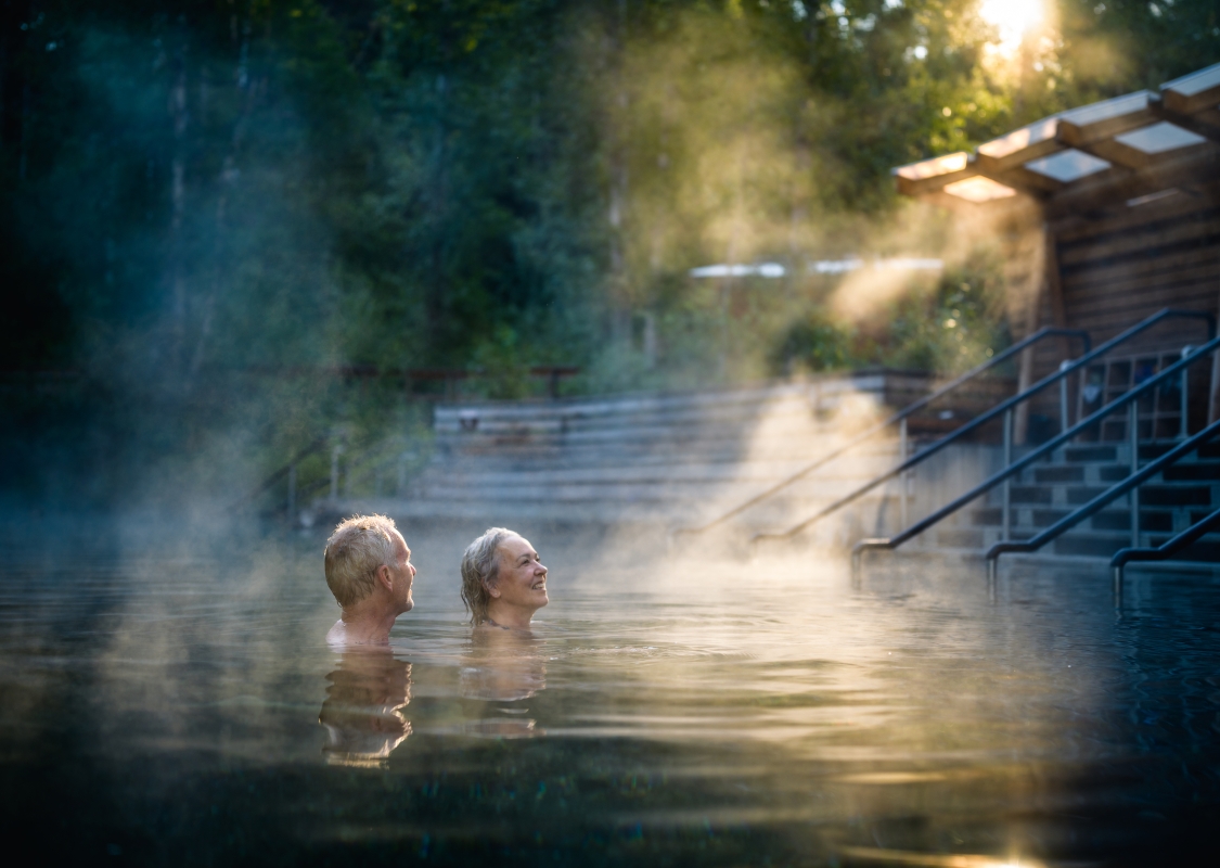 A couple soaks in hot springs with steam rising from the pools as sun peeks through the trees.