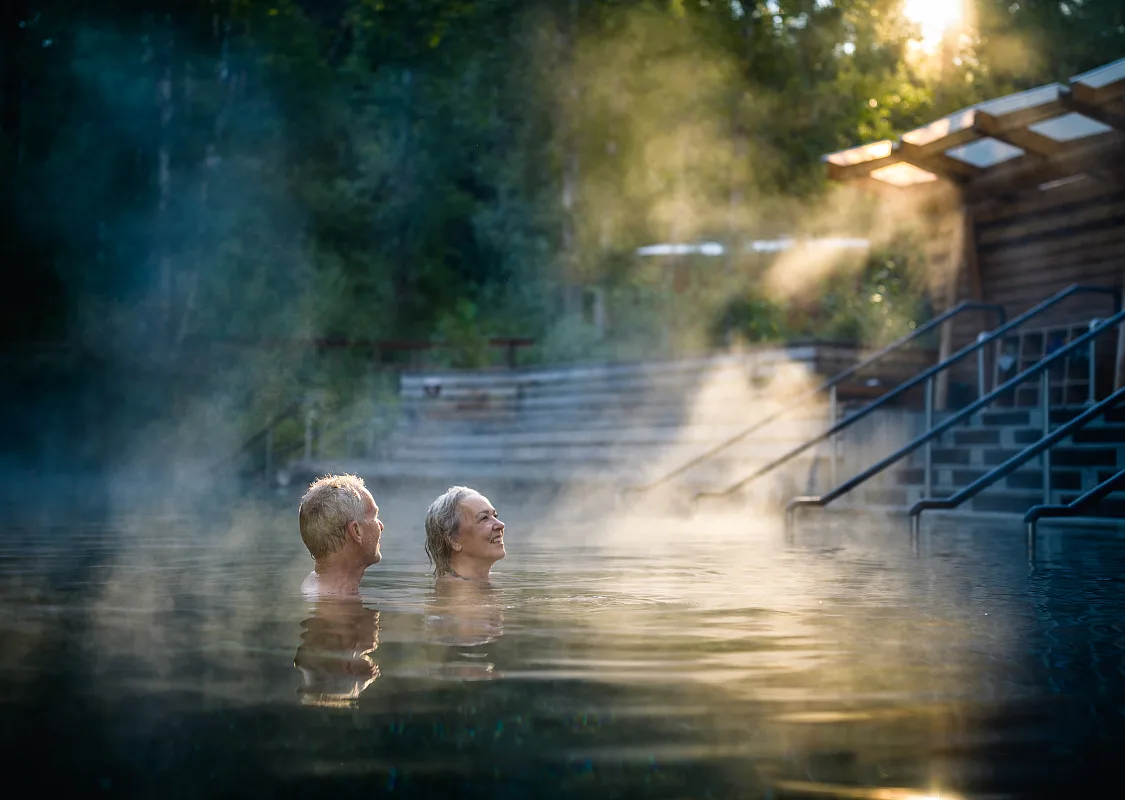 A couple soaks in hot springs with steam rising from the pools as sun peeks through the trees.