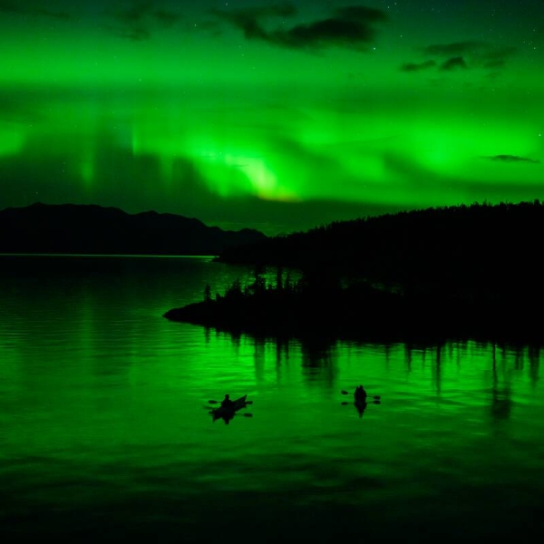 Two people kayak at night under the green glow of the Northern Lights.