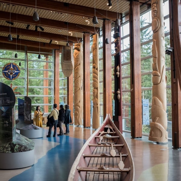 An Indigenous guide speaks to two visitors in an open gallery with cedar beams and tons of natural light. A wooden canoe sits in the foreground to the right, with totem poles and other carvings behind.
