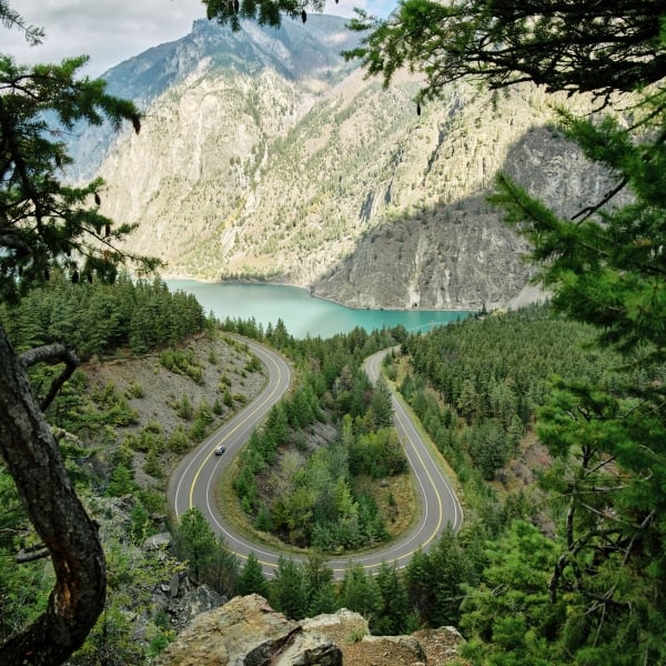 Vehicle travelling along a snaking highway with a turquoise lake and towering mountains in the background.