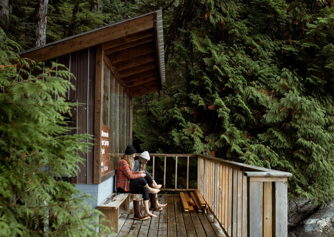 Two people sit near a wooden structure on a patio overlooking the forest.