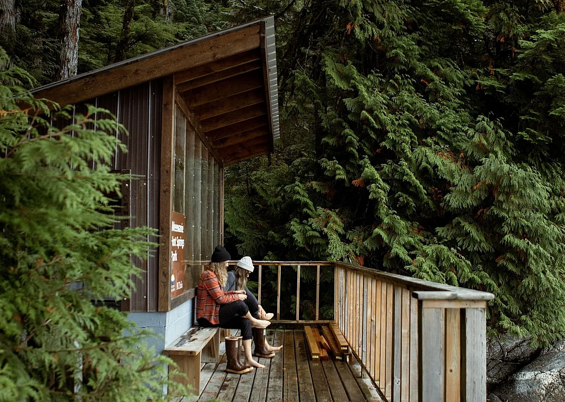 Two people sit near a wooden structure on a patio overlooking the forest.