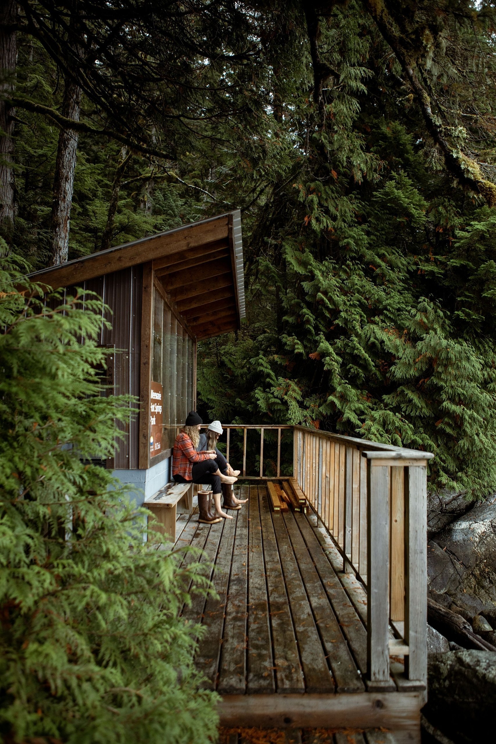 Two people sit near a wooden structure on a patio overlooking the forest.