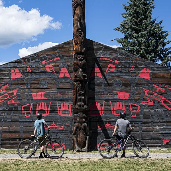 Two mountain bikers wheel their bikes past a longhouse with Indigenous artwork in 'Ksan.
