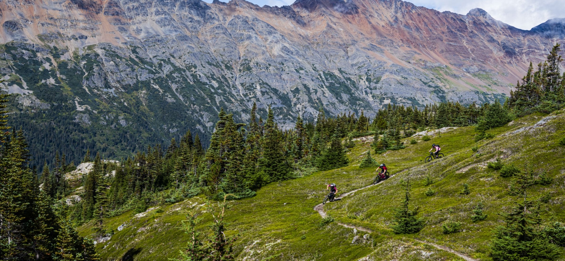 Los ciclistas se abren paso a través de un verde prado alpino con picos irregulares al fondo.