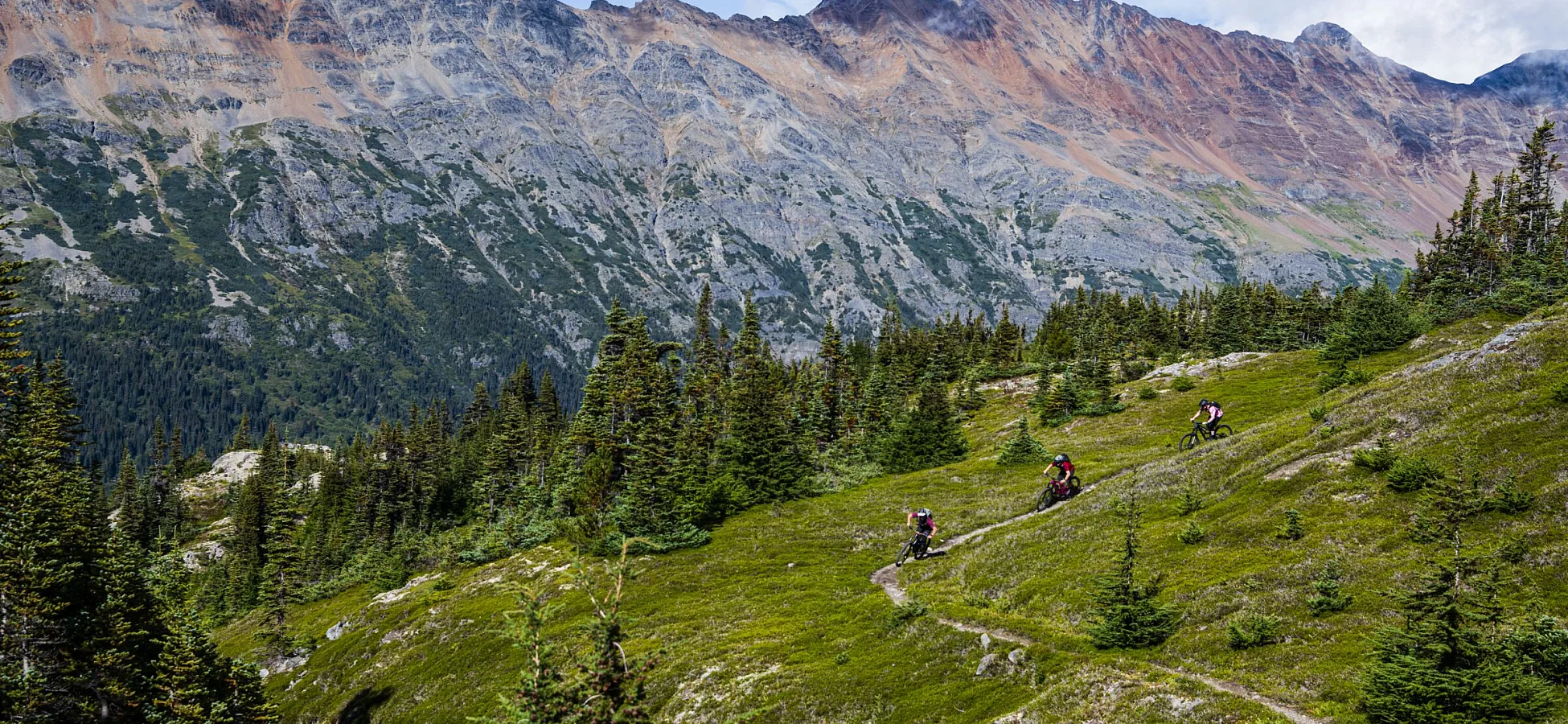 Riders weave their way through a green alpine meadow with jagged peaks in the background.