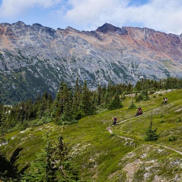 Riders weave their way through a green alpine meadow with jagged peaks in the background.