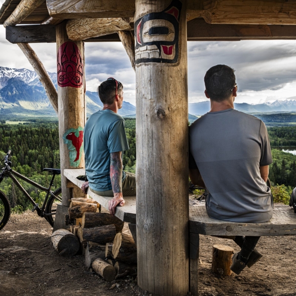 Dos ciclistas de montaña se sientan en un banco de madera tallado por indígenas mirando hacia un valle fluvial con montañas nevadas al fondo.
