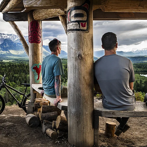 Two mountain bikers sit at an Indigenous-carved wooden bench looking out at a river valley with snow-capped mountains in the background.