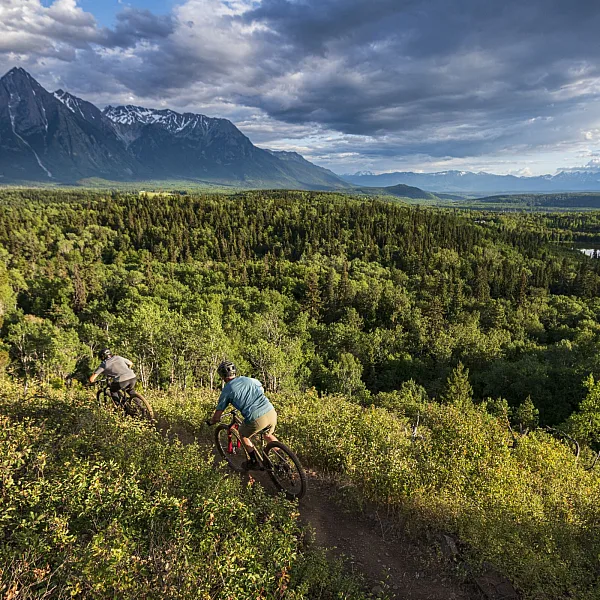 Bikers ride through a verdent trail with a river on the right-hand side and sharp mountains ahead.
