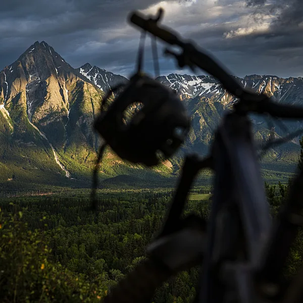 A mountain bike with dangling helmet sits in the foreground with a dramatic mountainscape with lush meadows in the background.