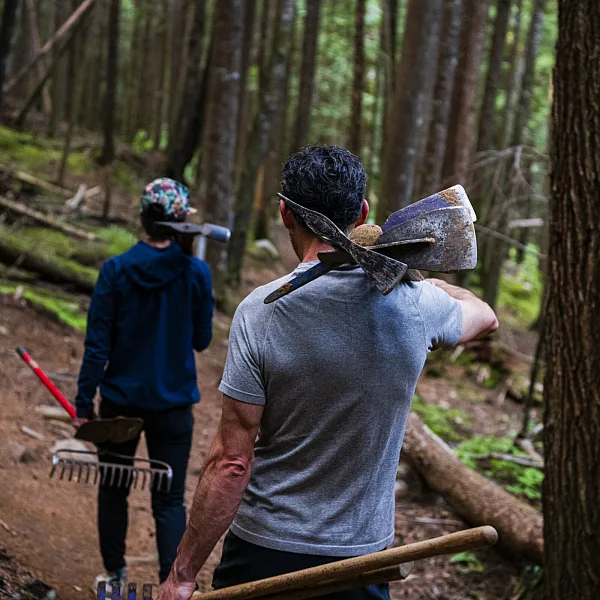 Two trail building volunteers walk along a trail with tools over their shoulders.