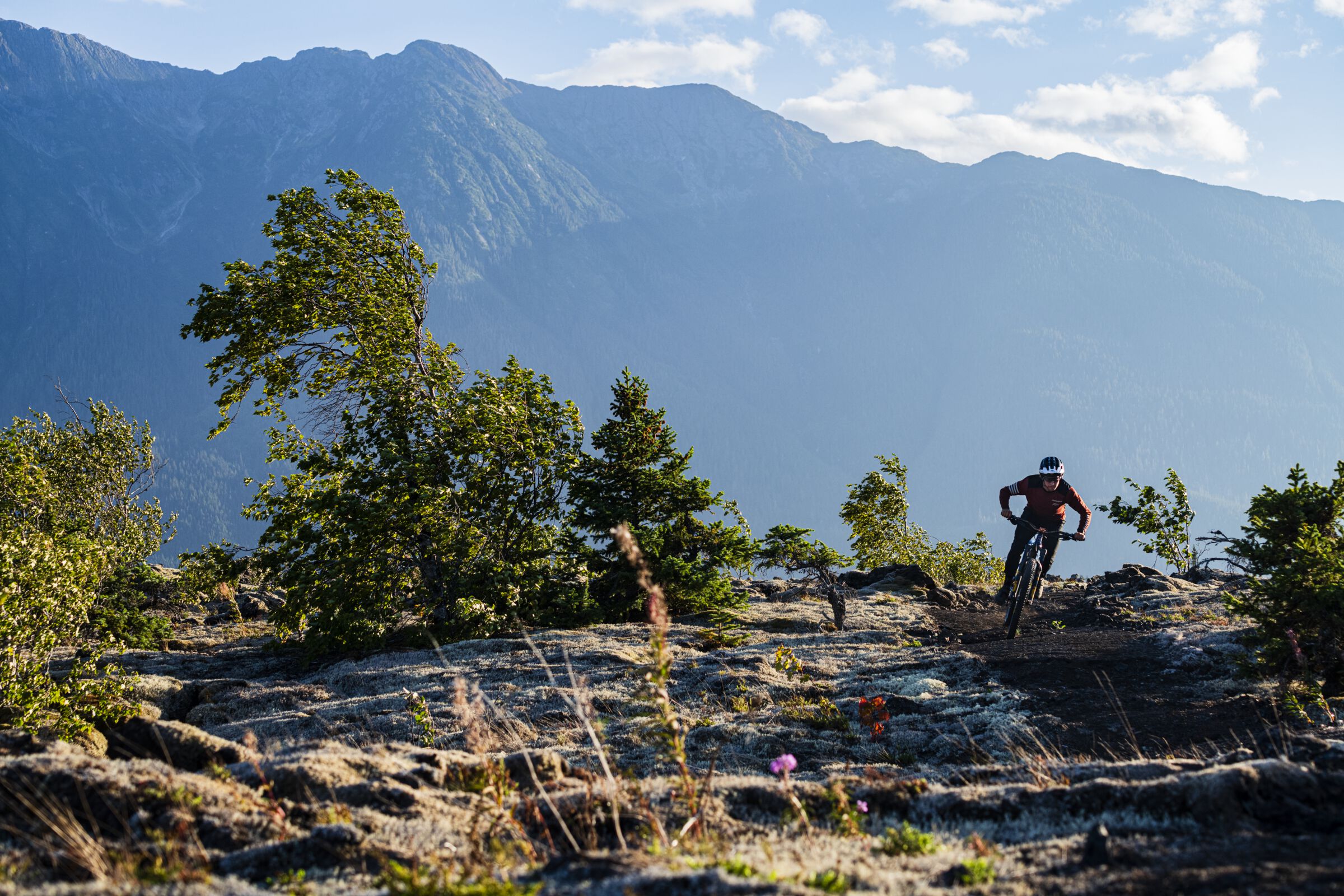 A mountain biker rides over a rocky outcropping with towering mountains in the backdrop.