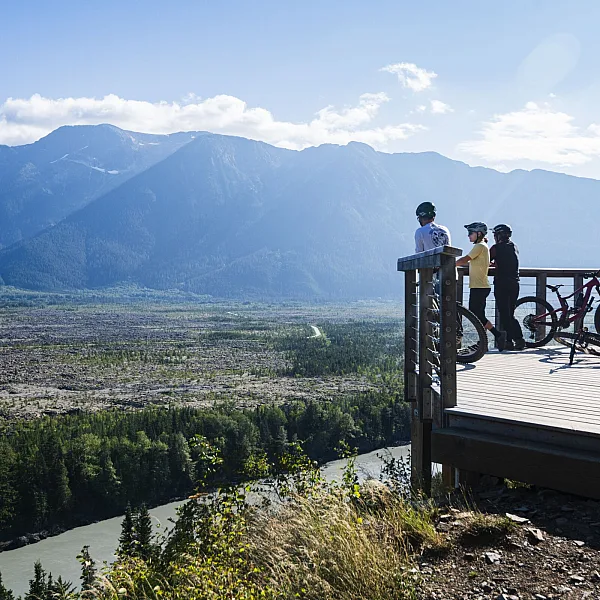 Two people on a wooden viewing platform, with bikes parked to the side, look out over a vast river valley.