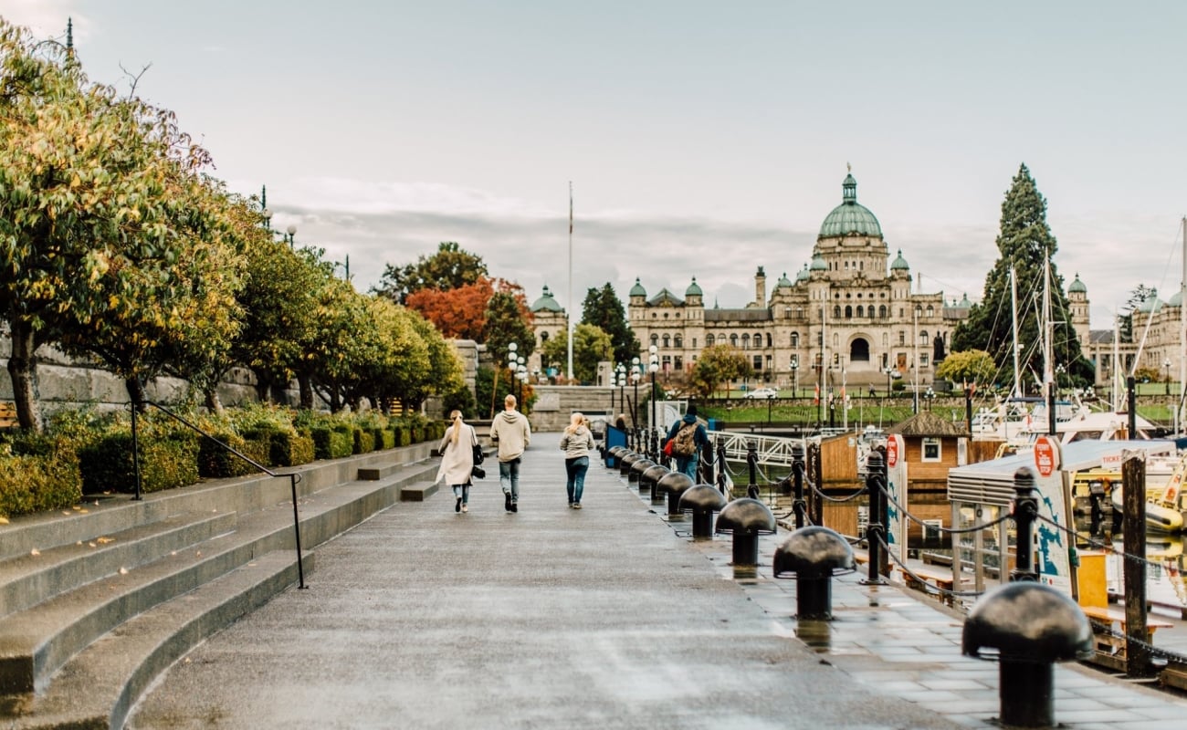 A family walks along the Inner Harbour in Victoria towards the Parliament Buildings. It's an overcast day and the path is wet. Boats sit in the docks by the harbour.
