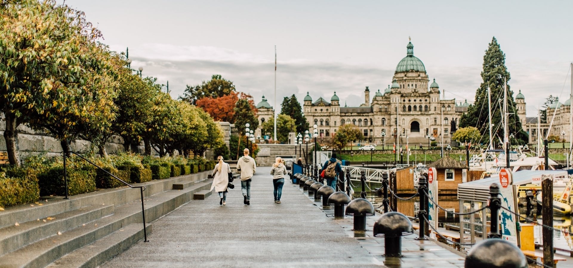 A family walks along the Inner Harbour in Victoria towards the Parliament Buildings. It's an overcast day and the path is wet. Boats sit in the docks by the harbour.