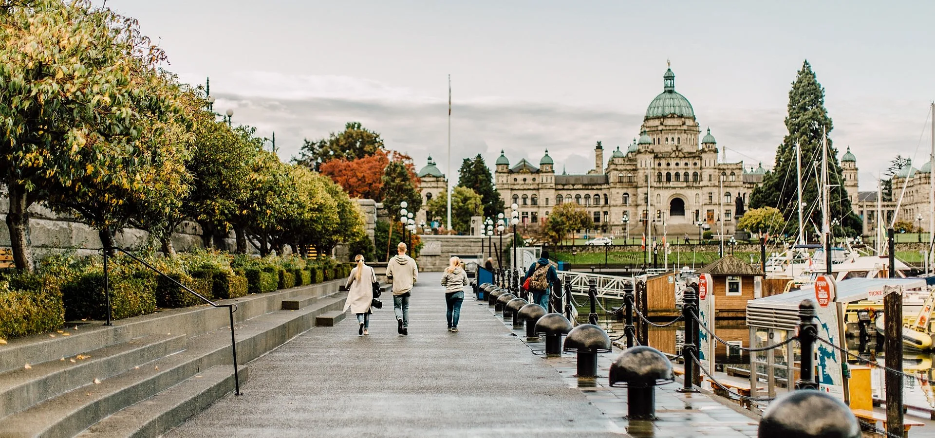 A family walks along the Inner Harbour in Victoria towards the Parliament Buildings. It's an overcast day and the path is wet. Boats sit in the docks by the harbour.