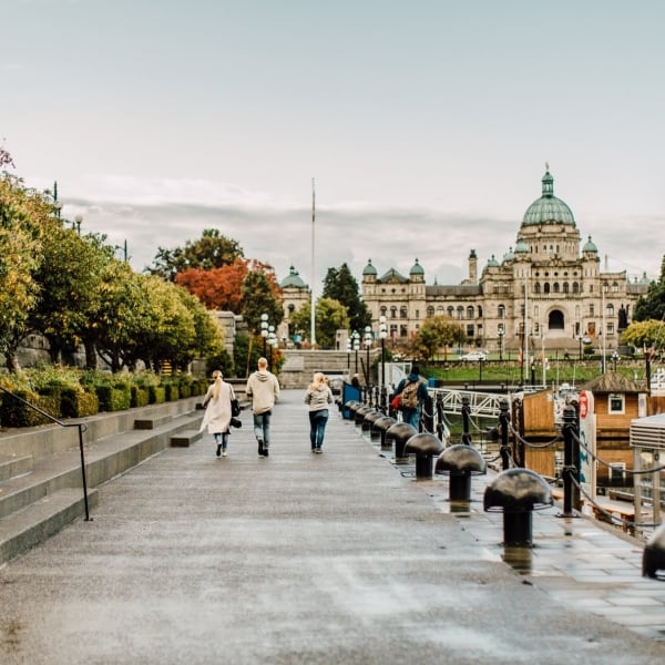 A family walks along the Inner Harbour in Victoria towards the Parliament Buildings. It's an overcast day and the path is wet. Boats sit in the docks by the harbour.