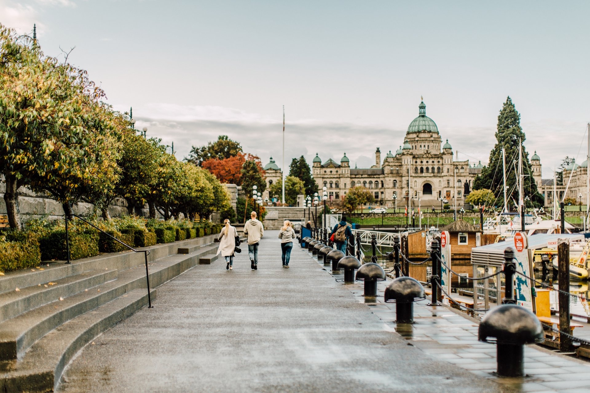 A family walks along the Inner Harbour in Victoria towards the Parliament Buildings. It's an overcast day and the path is wet. Boats sit in the docks by the harbour.