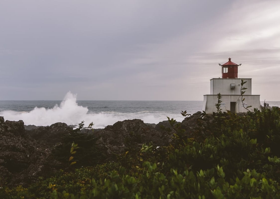 Sunset at Amphitrite Lighthouse at the Wild Pacific Trail in Ucluelet. The lighthouse has a red top level and two white levels. The spray from the ocean is crashing into the shoreline.