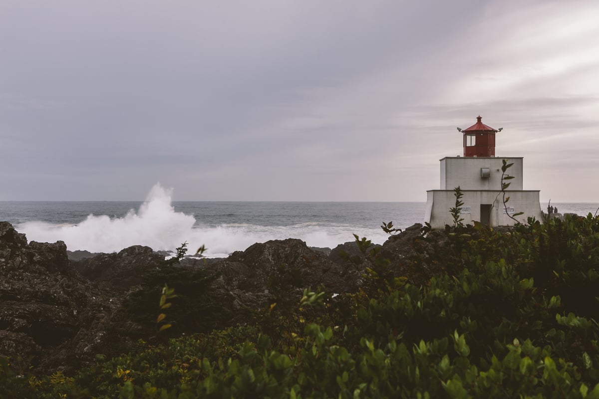 Sunset at Amphitrite Lighthouse at the Wild Pacific Trail in Ucluelet. The lighthouse has a red top level and two white levels. The spray from the ocean is crashing into the shoreline.