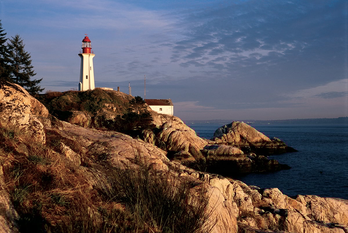 A red and white lighthouse sits on a rocky coastline at Lighthouse Park in West Vancouver. The sun is setting and the ocean is dark
