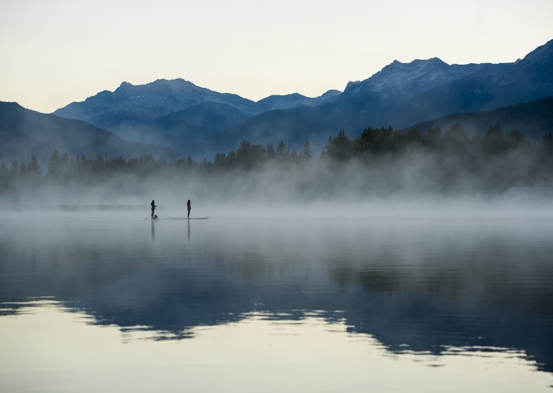 Two paddlers are out on a foggy lake in the morning. Mountains frame the background.