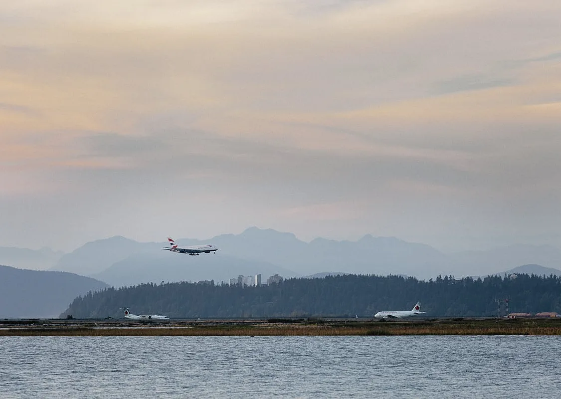 a plane lands at Vancouver International Airport. The photo is taken from across the Fraser River, and the North Shore Mountains are visible in the background.