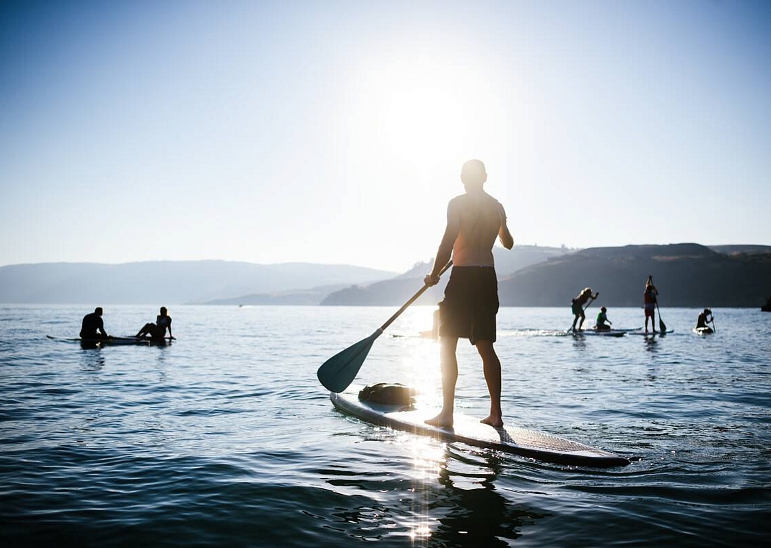 A person stands on a paddleboard in the foreground with half a dozen or so other paddleboarders in the distance. The sun is bright against the blue sky.