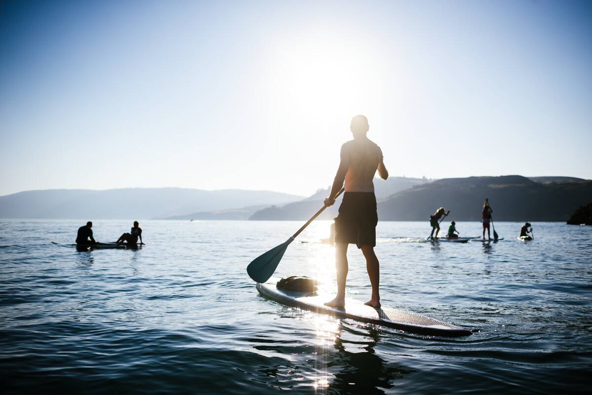 A person stands on a paddleboard in the foreground with half a dozen or so other paddleboarders in the distance. The sun is bright against the blue sky.