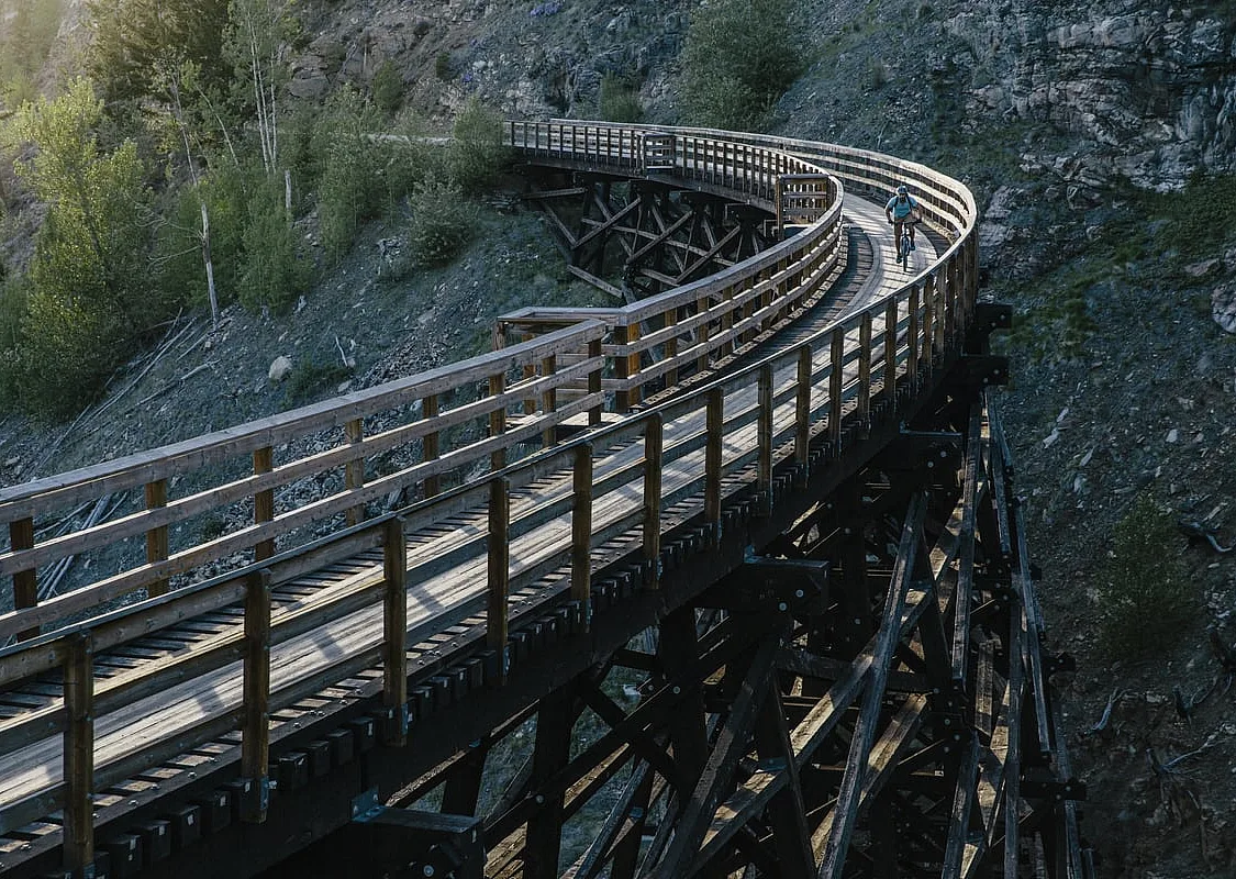 A wooden trestle bridge enters from the bottom left of the frame, and curves around and out the top left with a rocky bluff behind it