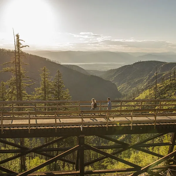 A person looks out from a trestle bridge in the Myra Canyon towards the mountain range. The sun is high in the sky and shines down on the green mountains below.