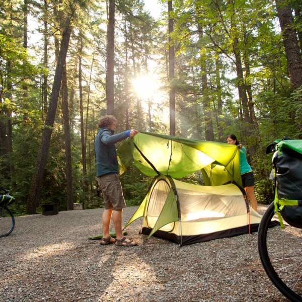 Couple setting up their tent at a campsite at a campsite surrounded by tall trees with sunlight shining through.